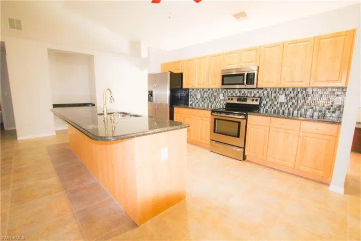 Kitchen with stainless steel appliances, light brown cabinets, dark stone countertops, an island with sink, and vaulted ceiling