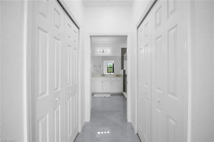 Hallway featuring white bi-fold doors and light-toned tile flooring, leading to a bathroom with a white vanity, wall-mounted lighting, and a framed mirror