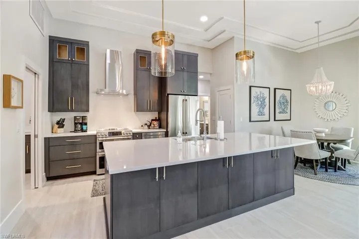 Kitchen with stainless steel appliances, wall chimney exhaust hood, decorative light fixtures, a large island with sink, and light stone counters