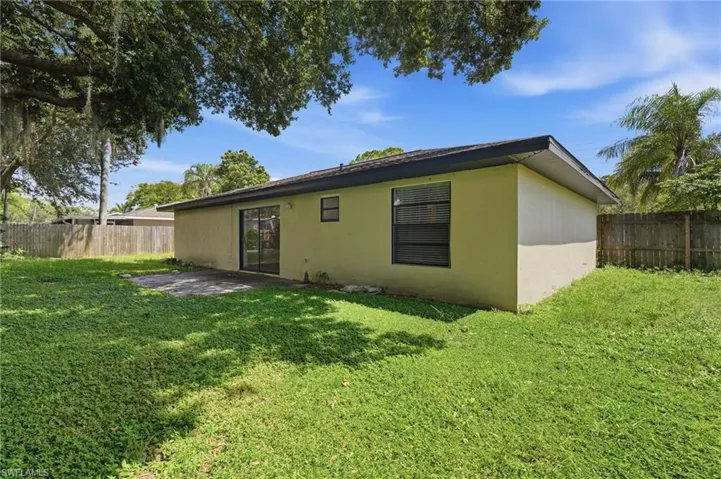 Spacious rear yard with a concrete patio, sliding glass doors, mature trees, and a privacy fence