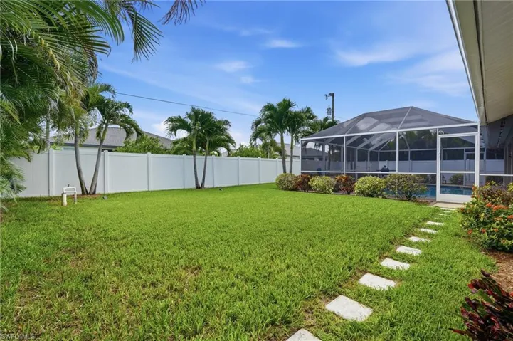 Expansive backyard featuring a green lawn, mature palm trees, a white privacy fence, a screened-in pool enclosure, and a stepping stone pathway