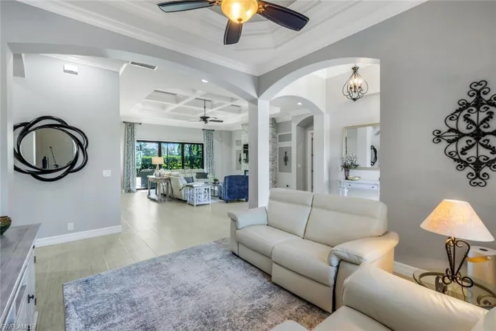 Featuring a high, coffered ceiling with white crown molding, the room is furnished with cream leather sofas, a light-colored area rug, and a wood-look floor in a light beige tone.