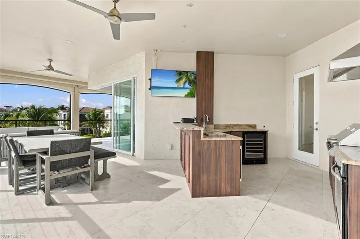Covered outdoor kitchen featuring stone countertops, built-in beverage refrigerator, wood-finish cabinetry, and stainless steel sink, complemented by large-format tile flooring and dual ceiling fans