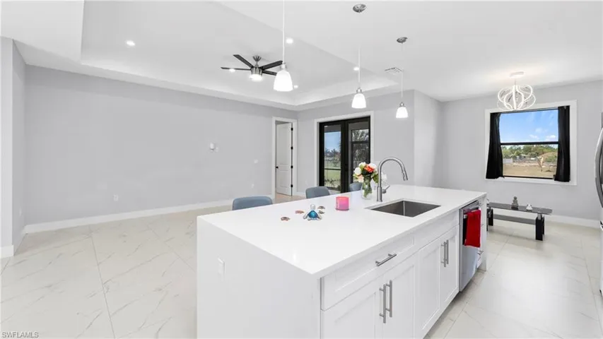 Kitchen featuring a kitchen island with sink, a raised ceiling, light stone countertops, hanging light fixtures, and white cabinetry