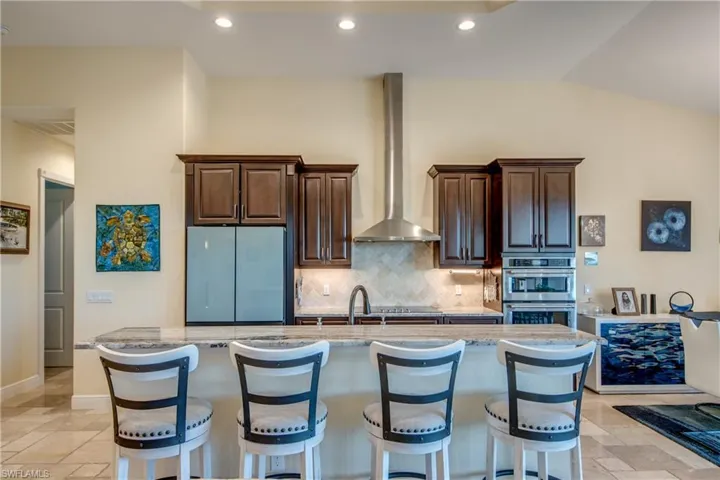 Kitchen with light stone counters, a breakfast bar area, tasteful backsplash, dark brown cabinetry, and wall chimney range hood