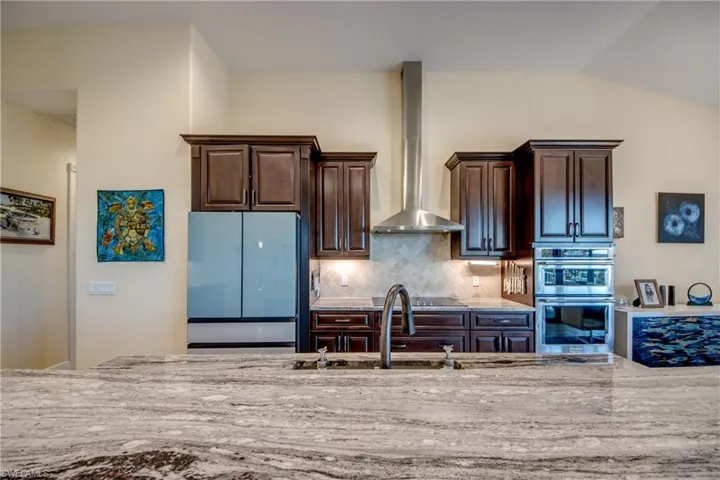 Kitchen with light stone counters, dark brown cabinets, fridge, wall chimney exhaust hood, and stainless steel double oven