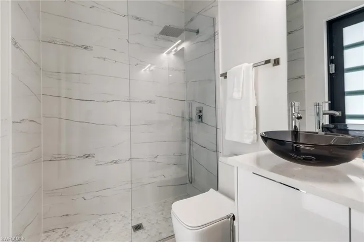 Contemporary bathroom featuring a clear glass shower enclosure with a rainfall shower head, white marble-style wall tiling, pebble-finish shower pan, black vessel sink, and a white vanity with a light-toned countertop