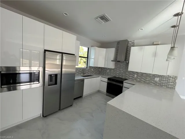 Kitchen featuring decorative backsplash, appliances with stainless steel finishes, wall chimney range hood, decorative light fixtures, and white cabinetry