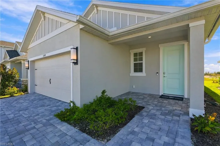 Single-story exterior featuring light-toned stucco siding, vertical gable trim, a paver driveway, covered front entryway, and wall-mounted exterior lighting fixtures