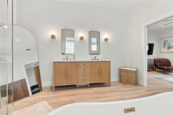 Dual-sink vanity with wood-finish cabinetry, countertop-integrated basins, brass-tone hardware, wall-mounted light fixtures, and wood-finish flooring