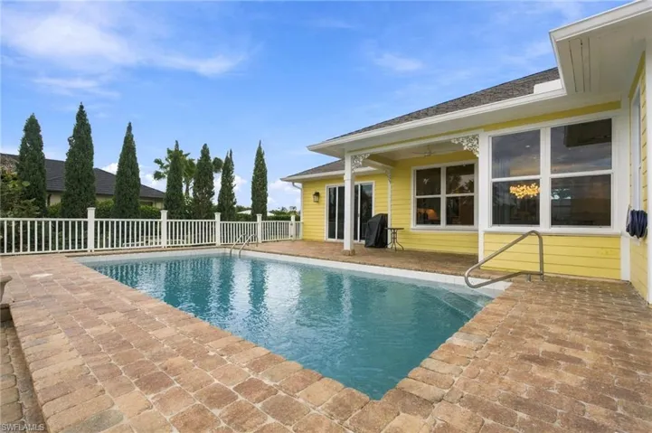In-ground swimming pool with stone-paver decking, covered patio, white picket fencing, and horizontal siding exterior