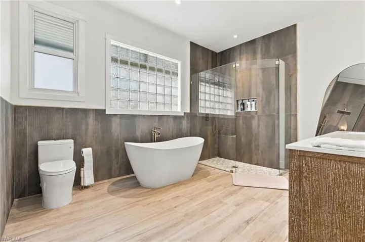 Freestanding soaking tub with floor-mounted faucet, walk-in glass-enclosed shower featuring built-in tiled niche, dark large-format wall tile, wood-finish flooring, and vanity with stone countertop
