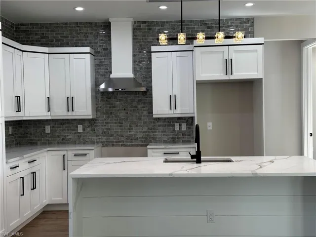 Kitchen featuring wall chimney exhaust hood, light stone counters, white cabinetry, and decorative light fixtures