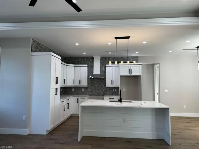 Kitchen with ceiling fan, an island with sink, wall chimney exhaust hood, and white cabinetry
