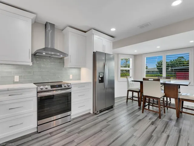 Kitchen with light hardwood / wood-style floors, wall chimney range hood, backsplash, white cabinetry, and appliances with stainless steel finishes