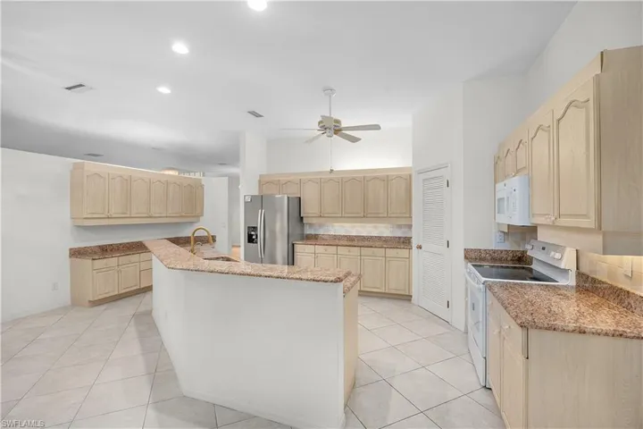 Kitchen with light brown cabinetry, white appliances, a kitchen island with sink, light tile patterned floors, and a ceiling fan