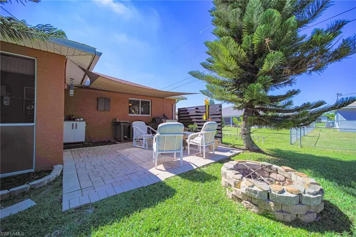 Rear exterior featuring a paved patio, stone-block fire pit, shaded area with a tension sail, and metal fencing