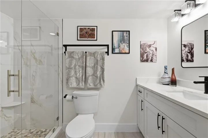 Shower with clear glass enclosure and light-toned tile, white vanity with integrated sink, black fixtures, rectangular mirror, and light wood-finish flooring