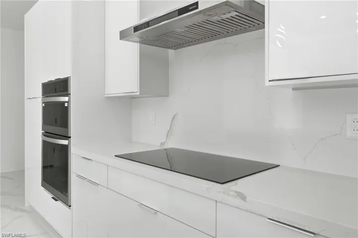 Integrated stainless steel double ovens, sleek induction cooktop, modern white cabinetry, stainless steel range hood, and continuous countertop and backsplash material