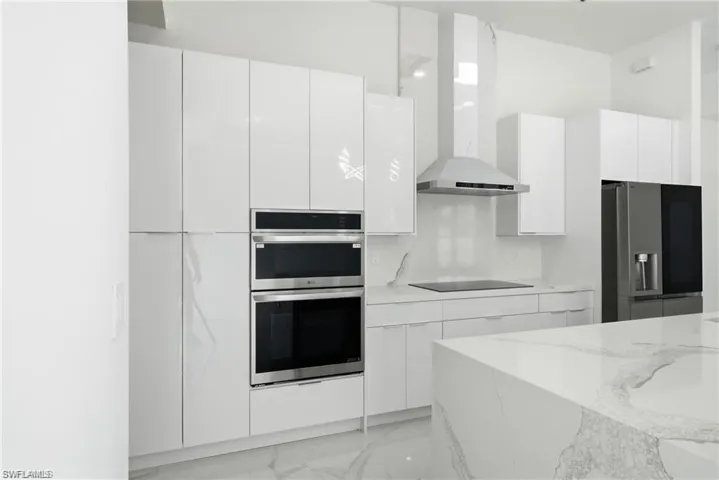 Contemporary kitchen featuring white flat-panel cabinetry, a built-in double oven, stainless steel range hood, and a spacious island with a stone countertop