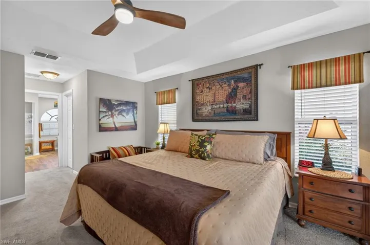 Bedroom featuring a tray ceiling with a ceiling fan, wall-to-wall carpeting, and a doorway opening to a bathroom with wood-finish flooring and a tiled arch window