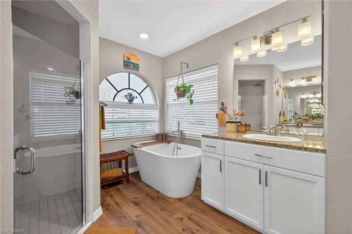 Bathroom featuring a freestanding soaking tub, wood-finish flooring, a glass-enclosed shower, a double vanity with a stone countertop, and an arched transom window