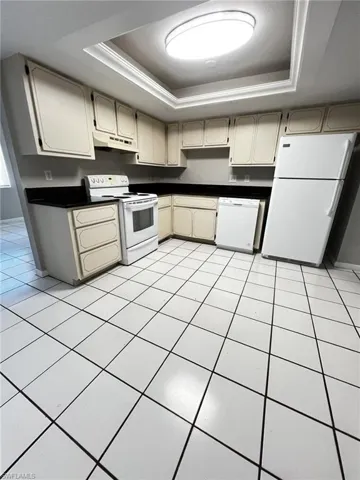 Kitchen featuring dark countertops, a tray ceiling, white appliances, light tile patterned floors, and cream cabinets