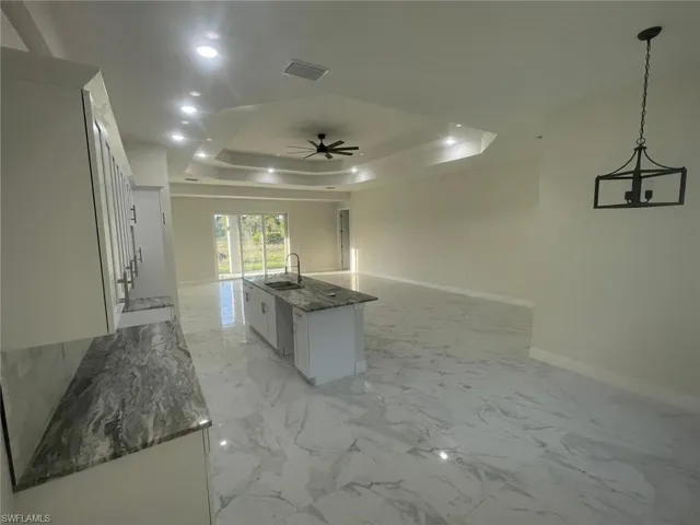 Kitchen with hanging light fixtures, a raised ceiling, a kitchen island with sink, white cabinets, and sink