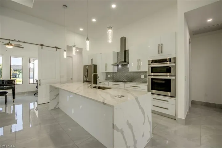 Kitchen with wall chimney range hood, sink, a kitchen island with sink, hanging light fixtures, and white cabinets