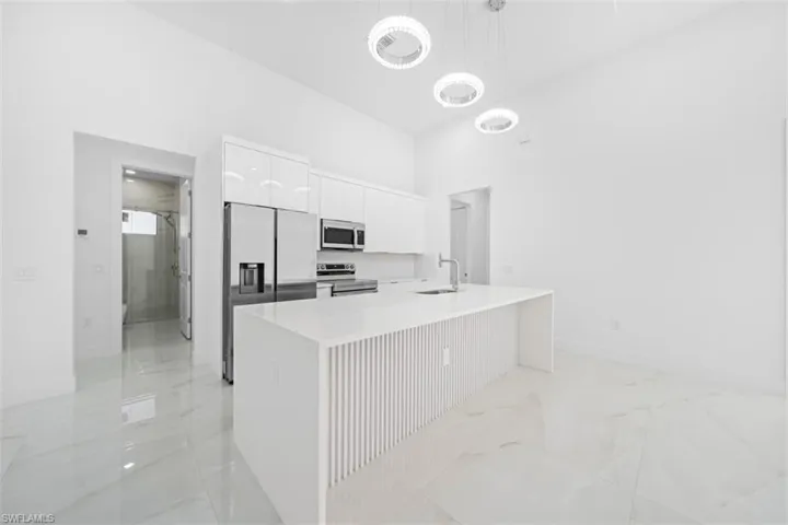 Contemporary kitchen featuring white gloss cabinetry, stainless steel appliances, a center island with a waterfall countertop, three pendant light fixtures, and large format polished tile flooring