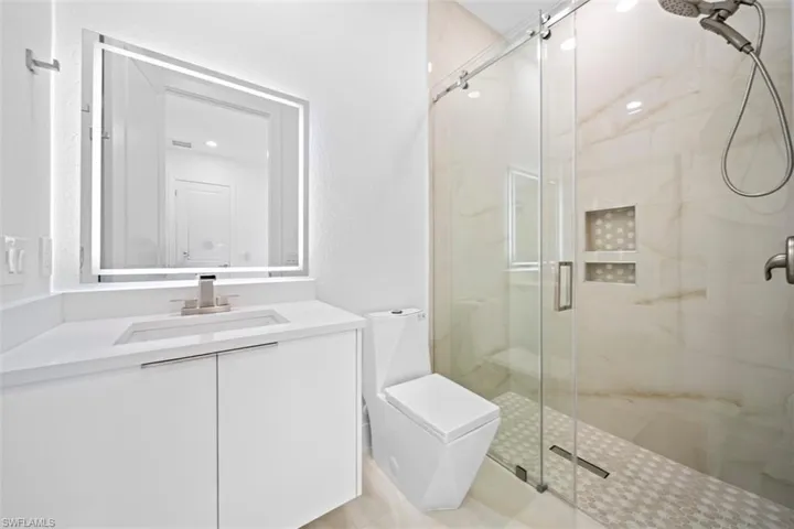 Modern bathroom featuring a frameless glass shower enclosure with a rain showerhead, a large vanity with an integrated sink and quartz countertop, a backlit mirror, and a contemporary toilet