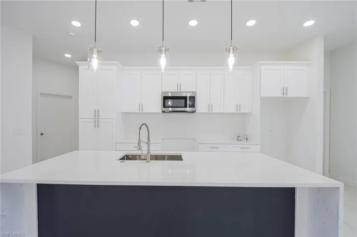 Kitchen with light stone counters, an island with sink, stainless steel microwave, pendant lighting, and two tone cabinets