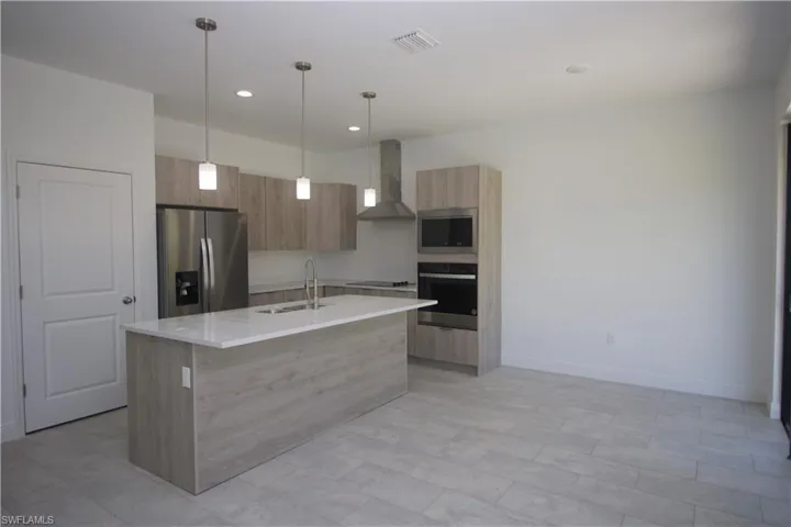 Kitchen featuring wall chimney exhaust hood, stainless steel appliances, sink, hanging light fixtures, and an island with sink