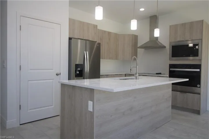 Kitchen featuring light stone counters, stainless steel appliances, a kitchen island with sink, wall chimney range hood, and decorative light fixtures