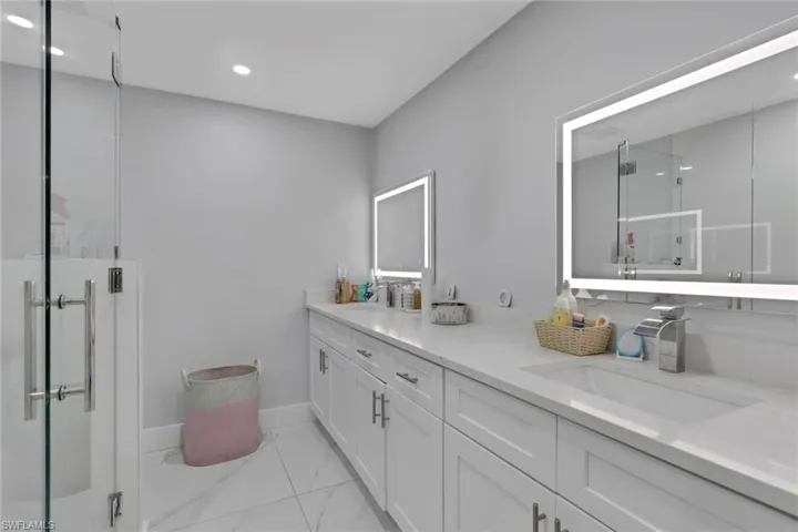 Dual vanity with integrated sinks, white cabinetry, illuminated mirrors, a glass-enclosed shower, and large format floor tiling