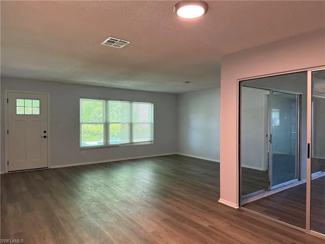 Spacious room featuring wood-finish flooring, a white paneled front door with window accents, and a large window with blinds