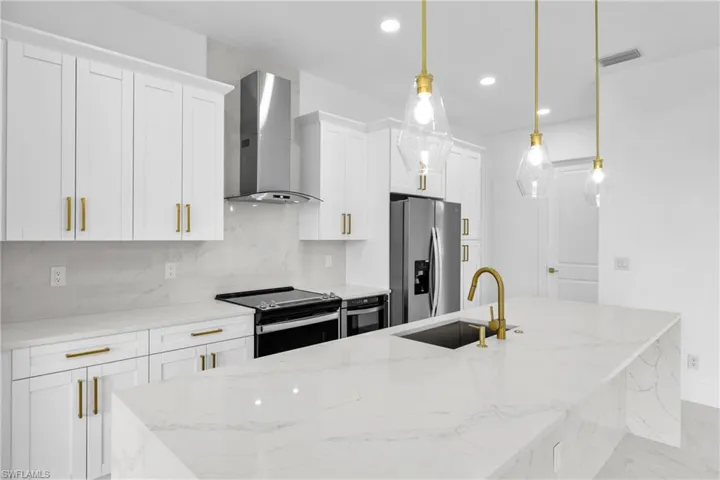 Kitchen featuring decorative light fixtures, light stone counters, tasteful backsplash, stainless steel appliances, and wall chimney range hood
