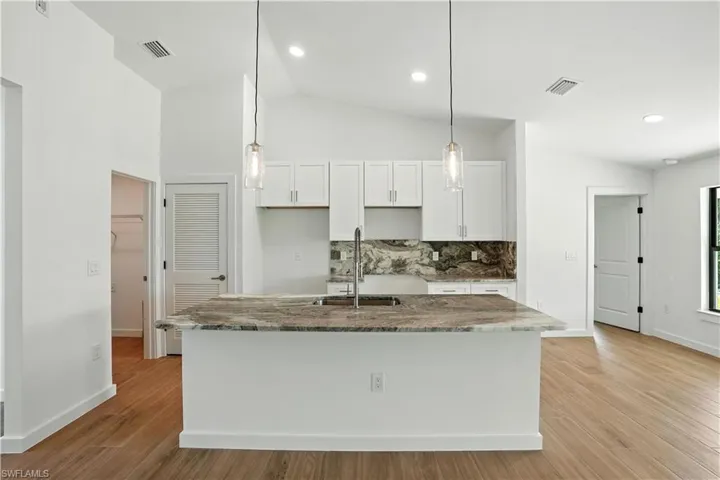 Kitchen featuring light stone counters, light wood finished floors, an island with sink, white cabinets, and backsplash