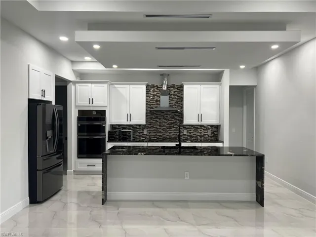 Kitchen with dark stone counters, recessed lighting, refrigerator with ice dispenser, white cabinets, and a kitchen island with sink