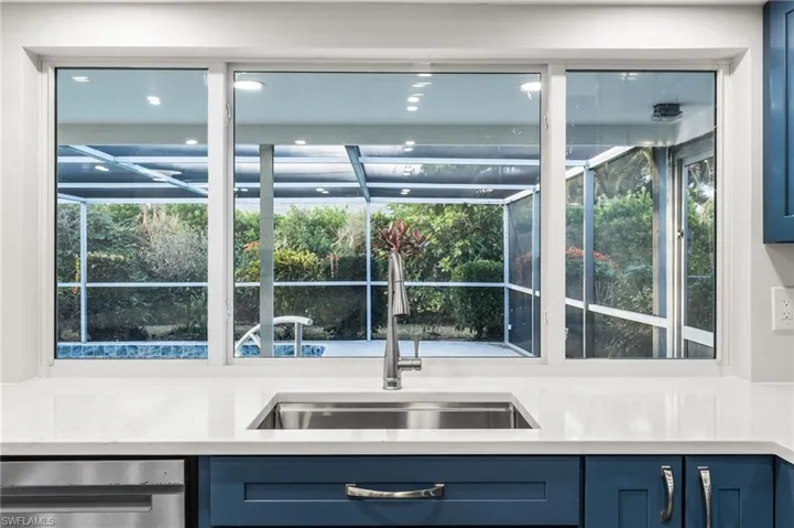 Kitchen with blue cabinets, light stone counters, and stainless steel dishwasher and a beautiful view of the pool and tropical landscaping.