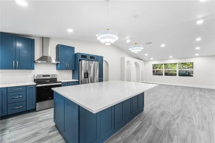 Kitchen with blue cabinetry, appliances with stainless steel finishes, vaulted ceiling, recessed lighting, and wall chimney exhaust hood