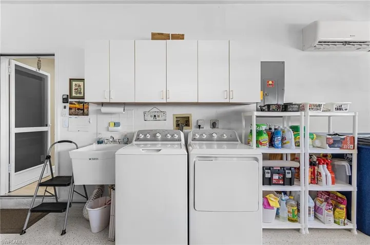 Laundry area with electric panel, cabinet space, a wall unit AC, washing machine and clothes dryer, and dark aggregate flooring