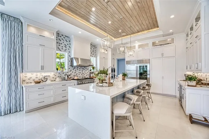 Kitchen with a raised ceiling, a center island with sink, built in fridge, wall chimney range hood, and ornamental molding