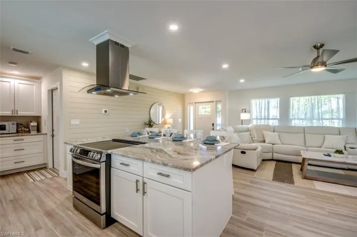 Open concept living area featuring a kitchen with a large island, stainless steel appliances, and light-colored cabinetry, opening into a living space with a ceiling fan and large windows