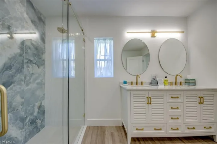 Bathroom featuring a dual vanity with gold hardware, two oval mirrors, and a shower with blue marble-style tile