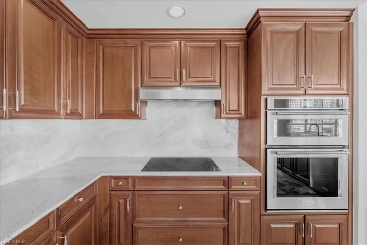 Kitchen featuring double oven, light stone counters, brown cabinetry, black electric stovetop, and under cabinet range hood