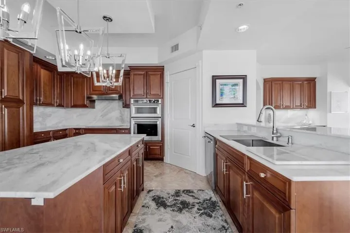 Kitchen featuring light stone counters, hanging light fixtures, appliances with stainless steel finishes, brown cabinetry, and a chandelier