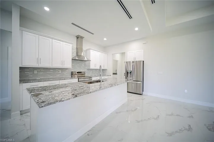 Kitchen with white cabinets, light stone countertops, stainless steel appliances, wall chimney range hood, and an island with sink