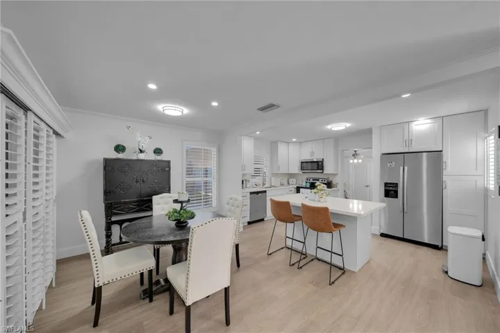 Open-concept kitchen and dining area featuring light wood-finish flooring, white cabinetry, stainless steel appliances, a kitchen island with seating, and plantation shutters