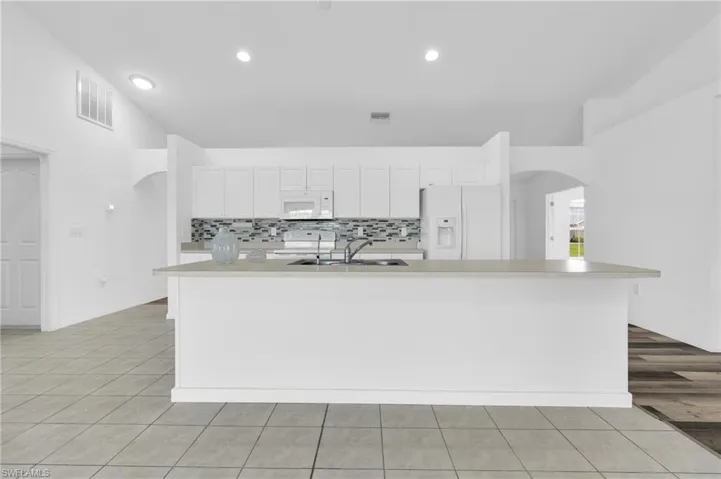 Kitchen featuring white appliances, arched walkways, light countertops, white cabinetry, and a center island with sink